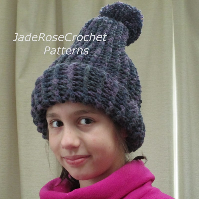 Pode incluir: Um gorro de croch&ecirc; roxo e cinza com um pompom no topo. O gorro est&aacute; sendo usado por uma pessoa com cabelo castanho e est&aacute; puxado para baixo sobre a testa.