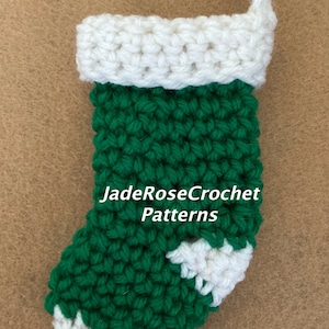 May include: A small green and white crocheted Christmas stocking with a white cuff. The stocking has a white crocheted design on the toe. The text "JadeRoseCrochet Patterns" is visible on the stocking.