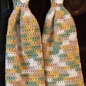 May include: Two crocheted kitchen towels with a scalloped edge, featuring a multi-coloured pattern of green, brown, white, and yellow. The towels have a button on each loop.