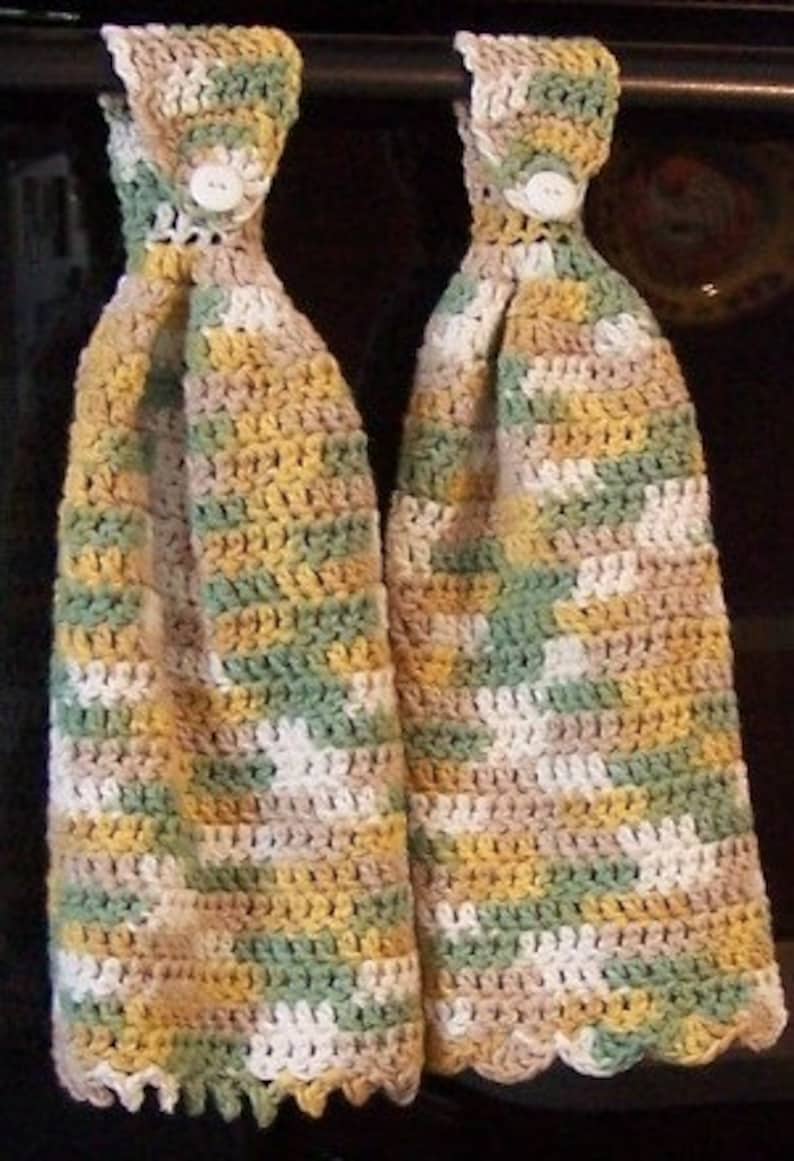 May include: Two crocheted kitchen towels with a scalloped edge, featuring a multi-coloured pattern of green, brown, white, and yellow. The towels have a button on each loop.