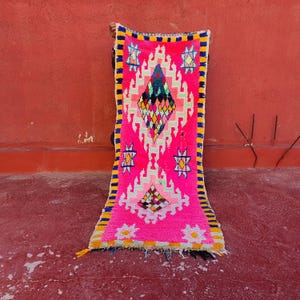 Peut inclure: Un tapis rose vif avec des motifs géométriques. Le tapis présente un motif central en losange de différentes couleurs, entouré de formes blanches, vertes et jaunes. Les bords sont bordés de rayures jaunes et bleues, et le tapis a un fond frangé.