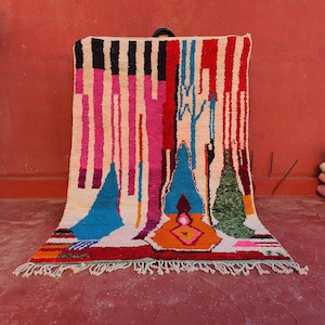 May include: A colorful, hand-woven rug with a geometric design. The rug features vertical stripes in red, black, pink, and blue, with abstract shapes in turquoise, orange, and green. The rug has a cream-colored background and a fringed edge.