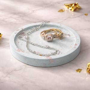 Op de afbeelding: Een gouden ring met een vierkante, heldere steen en een bijpassende band ligt op een lichtblauwe, ronde tray. Een zilveren ketting en gouden folie accenten zijn ook aanwezig. De tray staat op een marmeren oppervlak.
