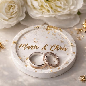 Könnte beinhalten: Ein weißer, runder Ringhalter mit Goldflocken und den Namen "Marie & Chris" in goldener Schrift. Zwei Eheringe liegen auf dem Halter, mit weißen Blumen im Hintergrund. Romantisches Hochzeitsthema.