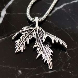 May include: Silver-toned leaf pendant necklace. The pendant is a detailed, stylized leaf design with intricate veining. The necklace chain is a small, linked design. The pendant and chain are set against a dark, marbled background.