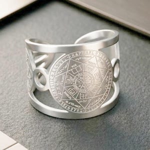 Op de afbeelding: Zilverkleurige manchetarmband met een ingewikkeld ontwerp. De armband heeft een centraal cirkelvormig embleem met gedetailleerde gravures en tekst, omgeven door opengewerkte patronen. Het metaal heeft een gepolijste afwerking. Een stijlvol accessoire.