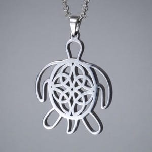 Könnte beinhalten: Ein silberfarbener Metallanhänger in Form einer Meeresschildkröte, der an einer passenden Gliederkette hängt. Der Panzer der Schildkröte weist ein kompliziertes, durchbrochenes Design aus ineinandergreifenden Kreismustern auf.