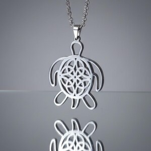 Op de afbeelding: Een zilverkleurige ketting met een schildpadhanger. De schildpadhanger heeft een open ontwerp met een Keltisch knoopmotief in het midden. De ketting is ook zilverkleurig en hangt aan de hanger.