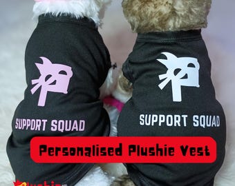 May include: Two plush toys wearing black vests with the text "SUPPORT SQUAD" and a graphic. The vests also have the text "Personalised Plushie Vest". One vest has pink graphics, the other white. The Plushie logo is at the bottom.