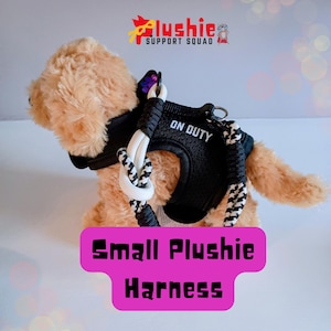May include: A soft toy dog wearing a black harness with the words "ON DUTY" in white. The harness is connected to a leash with black and white rope. The image also includes the text "Small Plushie Harness" on a pink background.