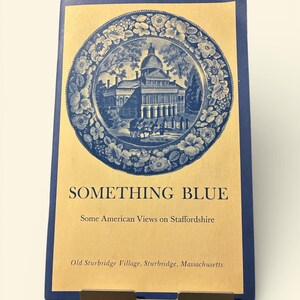 May include: A blue and tan booklet titled "SOMETHING BLUE" with an illustration of a building on a blue plate. The booklet is propped up by a dark metal stand. The text "Some American Views on Staffordshire" is below the title.