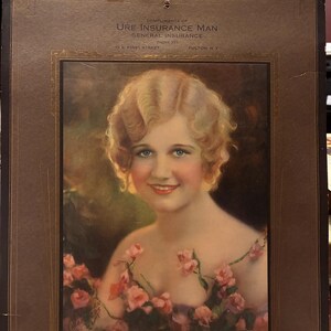 May include: Vintage print featuring a woman with blonde curls and pink flowers. The print is framed in a dark brown border and includes the text "URE INSURANCE MAN GENERAL INSURANCE" and the address "15 E. FIRST STREET FULTON, N.Y."