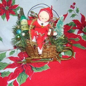 Könnte beinhalten: Eine rote und weiße Filzpuppe, die ein rotes und weißes Outfit trägt, sitzt in einem Weidenkorb mit einer Flasche Champagner, einem kleinen Weihnachtsbaum und Ornamenten. Die Puppe sitzt in einem Schlitten mit einem goldenen Metallrahmen.