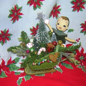 May include: A green sleigh filled with a miniature Christmas tree, candy canes, baubles, and a small doll dressed in green and holding a snowball. The sleigh is sitting on a red and white tablecloth with a poinsettia pattern.