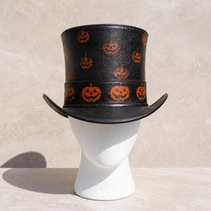 May include: A black top hat featuring a repeating pattern of orange jack-o'-lantern faces. The hat has a structured cylindrical crown and a curved brim, with a distinct band around the base also decorated with orange pumpkin designs.