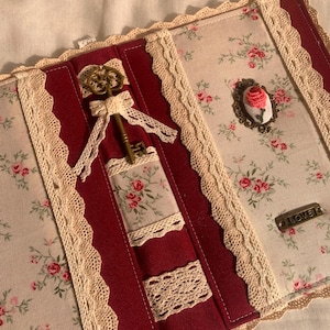 Pode incluir: Uma capa de livro em tecido com painéis alternados de tecido floral creme e tecido vermelho escuro sólido. É decorada com renda creme, uma chave cor de latão com um laço de renda, um broche de camafeu de rosa e uma etiqueta metálica com LOVE.