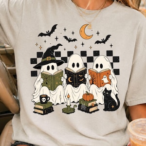 May include: A light beige short-sleeve t-shirt featuring a graphic print of three ghosts wearing witch hats and reading books. The design includes bats, a crescent moon, a black cat, a pumpkin, and a black and white checkered pattern background.