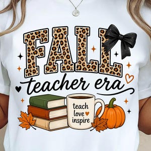 May include: A white t-shirt featuring the text FALL in a leopard print pattern with a black bow, and the text teacher era below. The design includes a stack of three books, a coffee mug with the text teach love inspire, and a pumpkin.