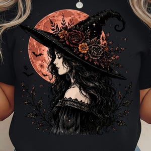 May include: A black t-shirt featuring a graphic illustration of a person with long dark hair wearing a wide-brimmed witch hat adorned with roses and leaves. A large orange moon, small flying bats, and delicate foliage surround the central figure.