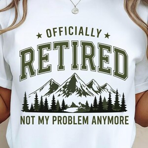Może przedstawiać: Biały t-shirt z krótkim rękawem z ciemnozielonym nadrukiem graficznym. Napis "OFFICIALLY RETIRED" znajduje się nad ilustracją gór i lasu sosnowego, a pod grafiką krajobrazu umieszczono napis "NOT MY PROBLEM ANYMORE".