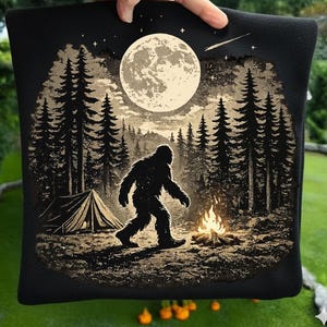 Pode incluir: Um item de tecido preto com uma estampa gráfica da silhueta do Bigfoot caminhando por uma floresta à noite. A cena inclui uma lua cheia grande, um céu estrelado com uma estrela cadente, uma barraca e uma fogueira brilhante em tons bege e preto.