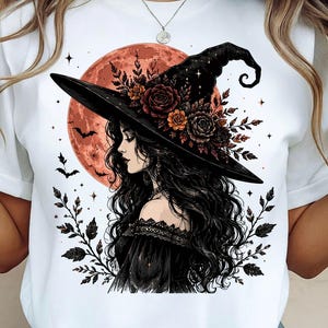 May include: A white t-shirt featuring a graphic illustration of a person with long dark hair wearing a black witch hat adorned with dark red and orange roses. The background shows a large orange moon, flying bats, and dark leafy branches.