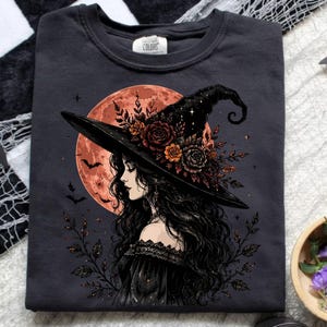 May include: A charcoal grey t-shirt featuring a graphic print of a person with long dark hair wearing a wide-brimmed witch hat adorned with dark roses and foliage. Behind the figure is a large orange moon with silhouettes of flying bats.