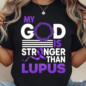Puede incluir: Camiseta negra con el texto "MY GOD IS STRONGER THAN LUPUS" en blanco y morado. El diseño incluye una corona de espinas morada y una cinta morada. La camiseta es de cuello redondo.