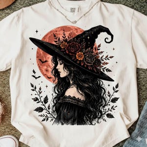 Cream-colored T-shirt mockup featuring a gothic witch with long flowing black hair, a black floral witch hat, a rusty orange moon, flying bats, and botanical details. The artwork is available as a high-resolution digital PNG download.