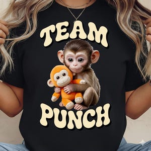 May include: Black t-shirt featuring a graphic of a monkey hugging an orange stuffed monkey. The text "TEAM PUNCH" is arched above and below the image in a retro font. The design is centered on the shirt.