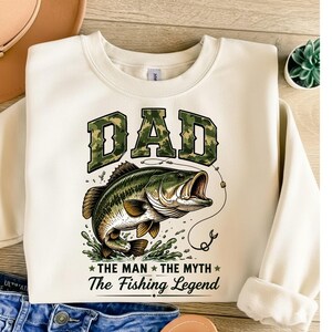 May include: Cream-colored sweatshirt with a fishing-themed graphic. The design features the word "DAD" in camouflage lettering above a detailed illustration of a largemouth bass. Below the fish, the text reads "THE MAN THE MYTH The Fishing Legend."