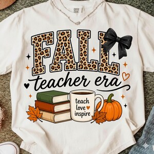 Puede incluir: Una camiseta color crema con la palabra FALL en estampado de leopardo y un lazo negro. Debajo, el texto dice teacher era. El dise&ntilde;o incluye una pila de tres libros, una taza de caf&eacute; con el texto teach love inspire y una calabaza.