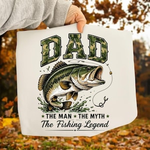 May include: White graphic print featuring a leaping bass fish with the text "DAD" in camouflage lettering. Below the fish, the text reads "THE MAN THE MYTH The Fishing Legend".