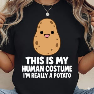 Pode incluir: Uma t-shirt preta com uma batata de desenho animado com um rosto sorridente e o texto branco: THIS IS MY HUMAN COSTUME I'M REALLY A POTATO. O gráfico da batata é castanho com manchas mais escuras e um contorno preto simples.