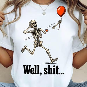 May include: A white t-shirt featuring a graphic of a skeleton running with one arm detached, holding a red balloon. The skeleton has a shocked expression, and the text "Well, shit..." is printed in black below the illustration.
