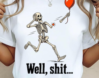 May include: A white t-shirt featuring a graphic of a skeleton running with one arm detached, holding a red balloon. The skeleton has a shocked expression, and the text "Well, shit..." is printed in black below the illustration.