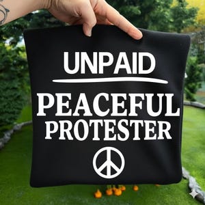 May include: Black tote bag featuring the white text "UNPAID PEACEFUL PROTESTER" and a peace symbol. The bag is held up against a green background.