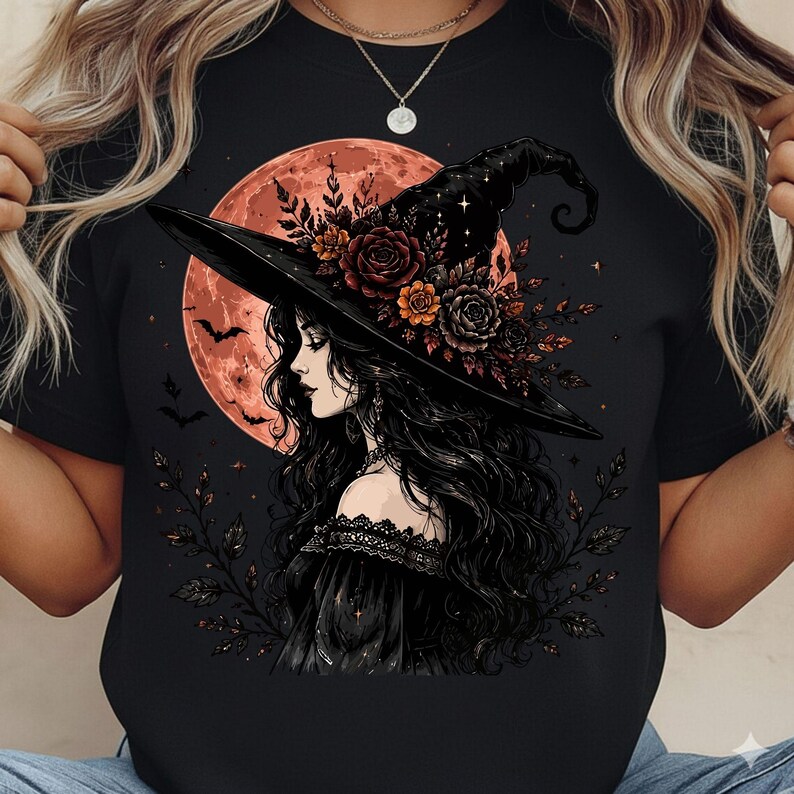 May include: A black t-shirt featuring a graphic illustration of a person with long dark hair wearing a wide-brimmed witch hat adorned with roses and leaves. A large orange moon, small flying bats, and delicate foliage surround the central figure.