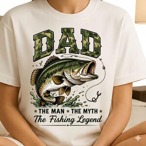 May include: White t-shirt featuring a large graphic of a leaping fish with an open mouth, a fishing hook, and the text "DAD" in camouflage lettering. Below the fish, the text reads "THE MAN THE MYTH The Fishing Legend."