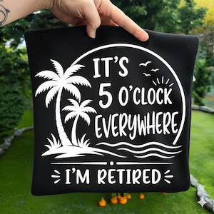 May include: Black square pillow with white text and graphics. The text reads "IT'S 5 O'CLOCK EVERYWHERE I'M RETIRED" with a palm tree and ocean waves design. The pillow is held up by a person.
