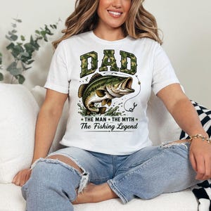 May include: White t-shirt with a graphic of a largemouth bass jumping out of water. The word "DAD" is in camouflage lettering above the fish, with the text "The Man The Myth The Fishing Legend" below.