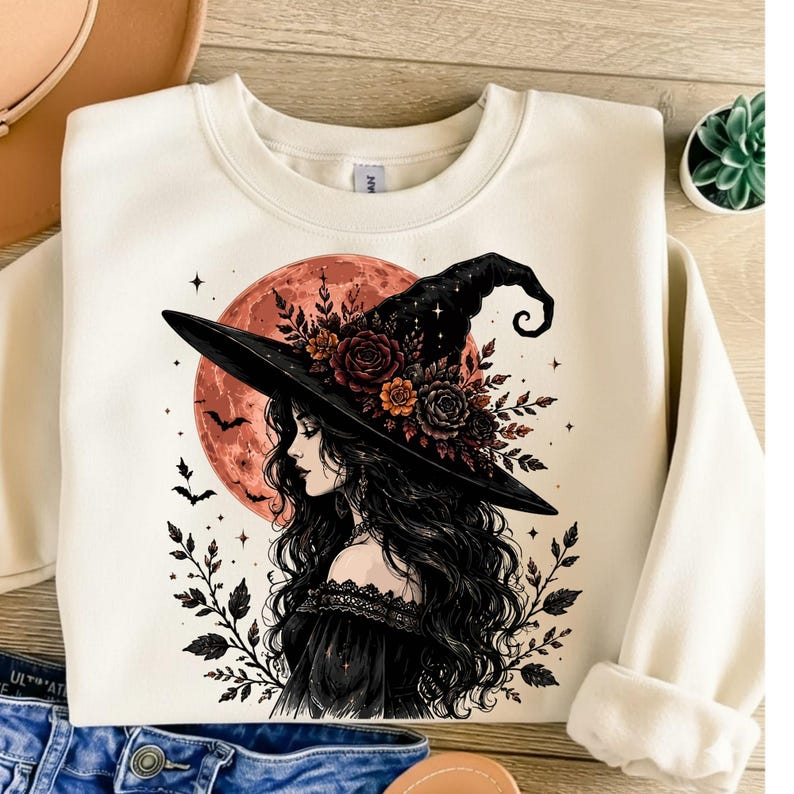 May include: A cream-coloured crewneck sweatshirt featuring a graphic print of a person with long dark hair wearing a black witch hat adorned with dark flowers. The design includes a large orange moon, small stars, flying bats, and leafy branches.