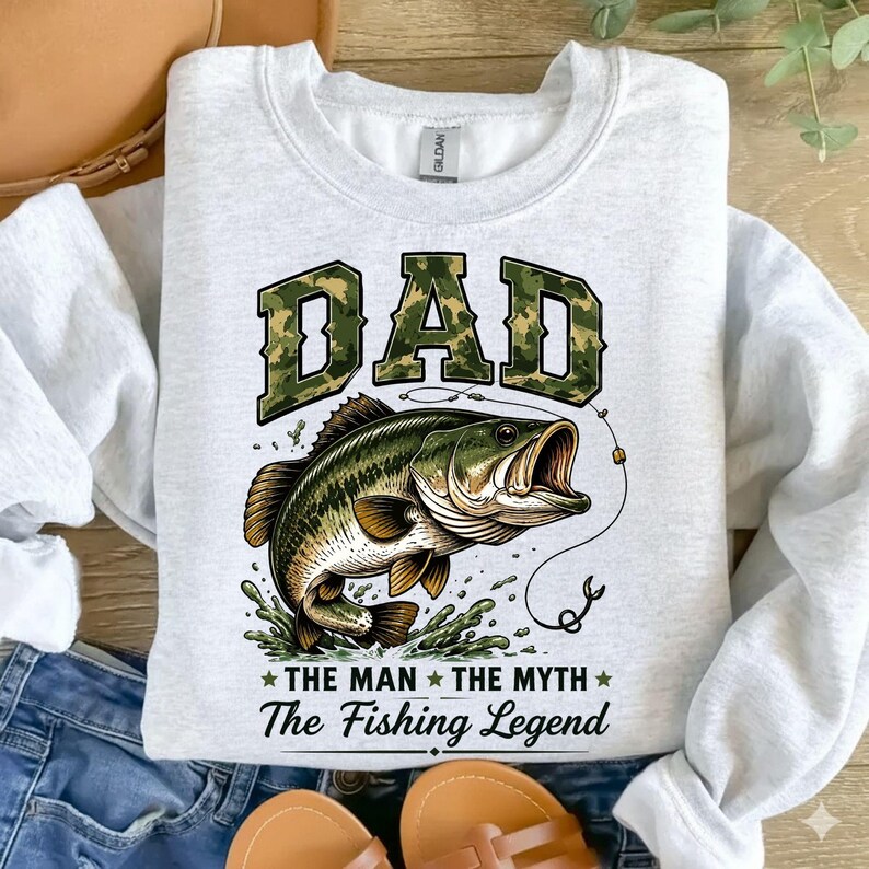 May include: Light gray sweatshirt with a graphic of a largemouth bass jumping out of water. The word "DAD" is in camouflage lettering above the fish. Below the fish, the text reads "THE MAN THE MYTH The Fishing Legend."
