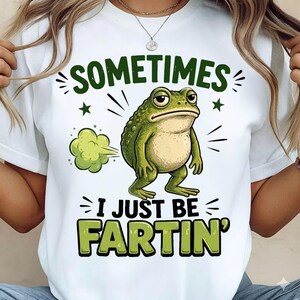 Könnte beinhalten: Weißes T-Shirt mit einer grünen Cartoon-Frosch-Grafik. Der Frosch hat einen grimmigen Gesichtsausdruck, und eine Gaswolke kommt aus seinem Hinterteil. Der Text auf dem Shirt lautet "SOMETIMES I JUST BE FARTIN'."