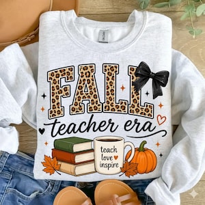 Puede incluir: Una sudadera gris claro de cuello redondo con el texto FALL en estampado de leopardo con un lazo negro, el texto teacher era, una pila de tres libros, una taza con el texto teach love inspire y un gr&aacute;fico de una calabaza naranja.