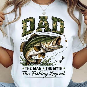 May include: White t-shirt with a graphic of a largemouth bass jumping out of water. The word "DAD" is in camouflage lettering above the fish, with the text "THE MAN THE MYTH The Fishing Legend" below.
