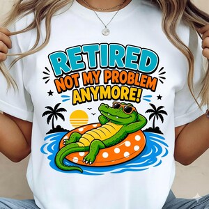 May include: White t-shirt with a graphic of a green alligator wearing sunglasses, relaxing on an orange and yellow inflatable ring in blue water. The text "RETIRED NOT MY PROBLEM ANYMORE!" is displayed above the alligator.