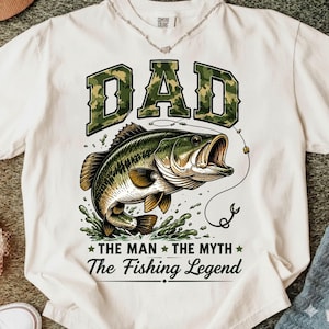 May include: Cream-colored t-shirt with a graphic of a largemouth bass and the word "DAD" in camouflage lettering. The text "THE MAN THE MYTH The Fishing Legend" is below the fish. A fishing-themed shirt.