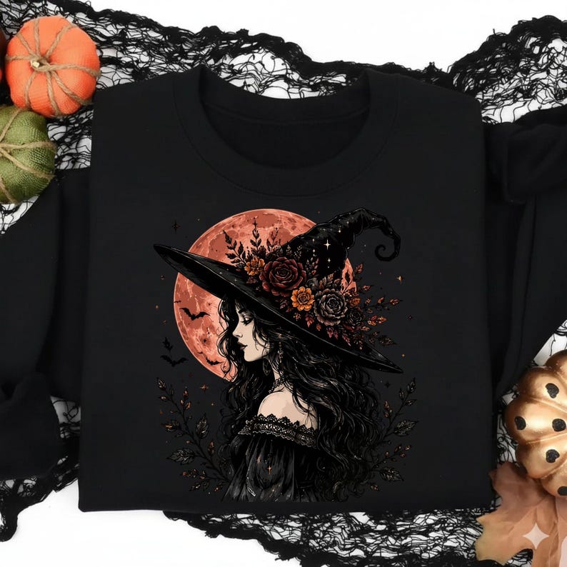 May include: A black crewneck sweatshirt featuring a graphic print of a person with long dark hair wearing a wide-brimmed witch hat adorned with dark red and orange roses. A large, glowing orange full moon and small bats appear in the background.