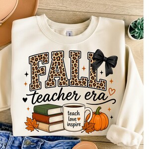 Puede incluir: Una sudadera de cuello redondo color crema con el texto FALL en estampado de leopardo con un lazo negro, y el texto teacher era debajo. El dise&ntilde;o incluye una pila de tres libros, una taza con el texto teach love inspire y una calabaza.